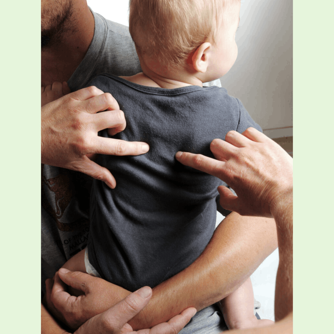Aceasta este o fotografie care ilustrează modul de lucru cu sugarii.  Imaginea: Un tată își ține bebelușul în brațe, cu spatele copilului orientat spre terapeut.  Acțiunea: Se observă mâinile terapeutului aplicând presiuni foarte ușoare cu degetele arătătoare pe spatele bebelușului, de o parte și de alta a coloanei vertebrale.  Context: Sugerează o metodă sigură și blândă, unde copilul rămâne în confortul brațelor părintelui în timpul tratamentului.